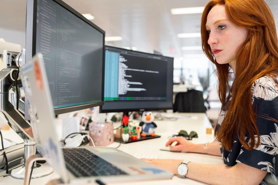 Female software developer with red hair looking at code on three monitors. | CaseGuard Female software developer with red hair looking at code on three monitors.